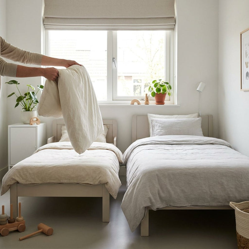 Ouder legt verschillende kinderdekbedden op een netjes opgemaakt kinderbed in een lichte slaapkamer, passend bij peuter- en eenpersoonsmaten.