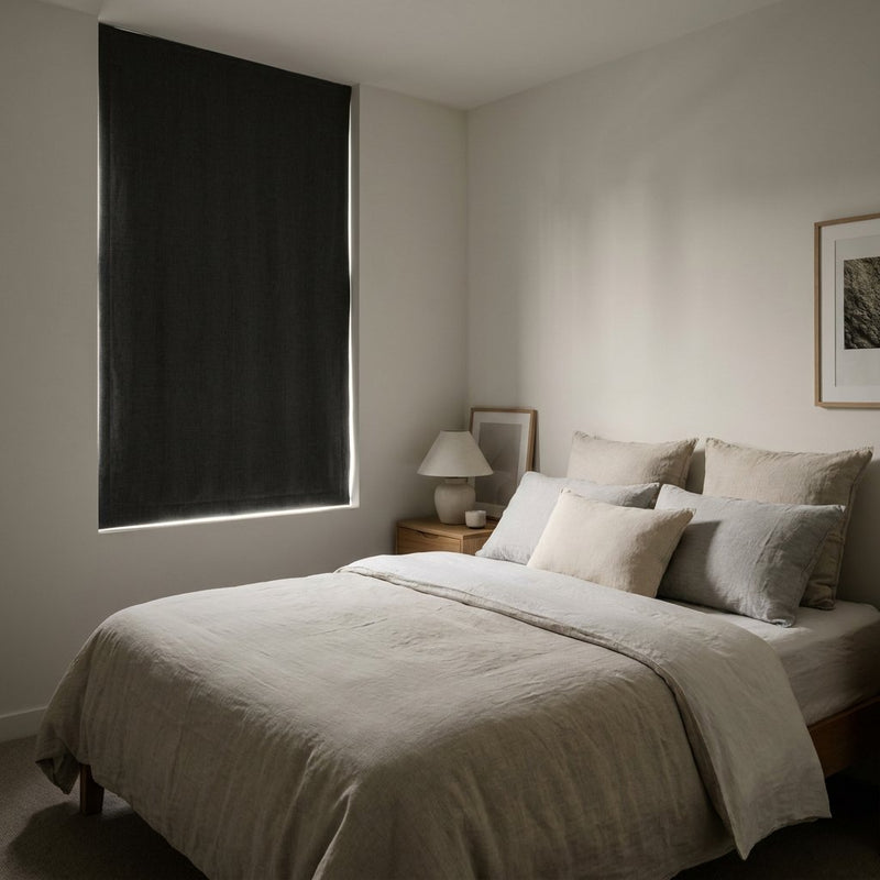 Donkere moderne slaapkamer met goed sluitende verduisterende gordijnen, zacht ochtendlicht langs het raam en een opgemaakt bed in neutrale tinten.