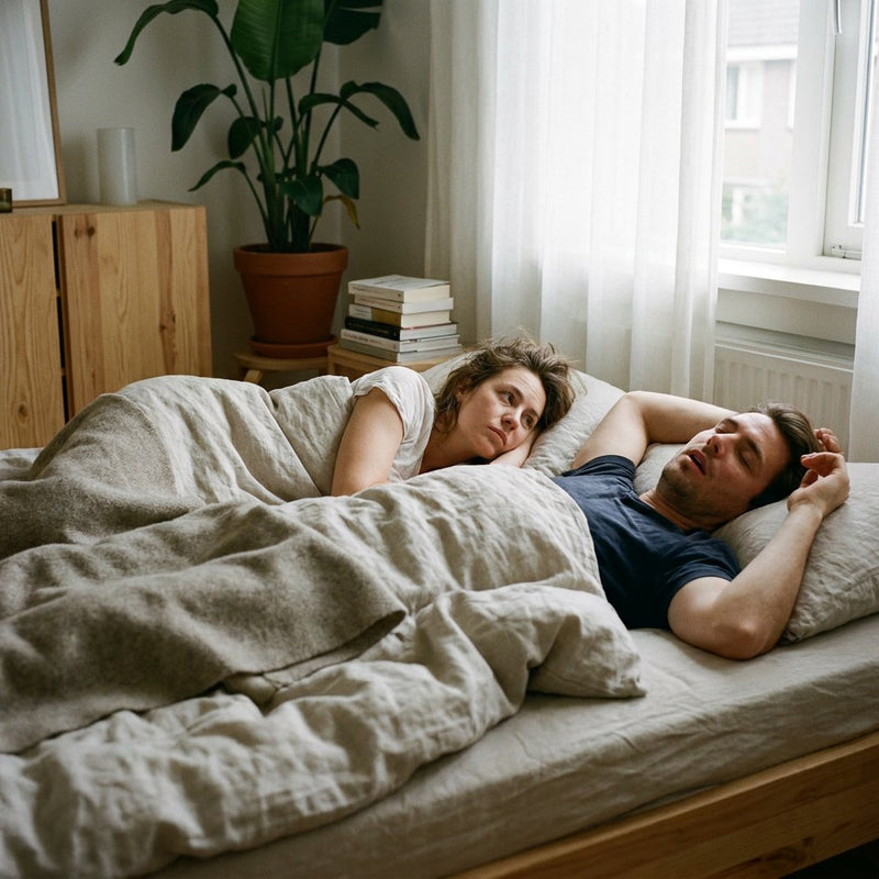 Vrouw ligt wakker naast een slapende, snurkende partner in een rustige moderne slaapkamer met zacht ochtendlicht en neutraal beddengoed.